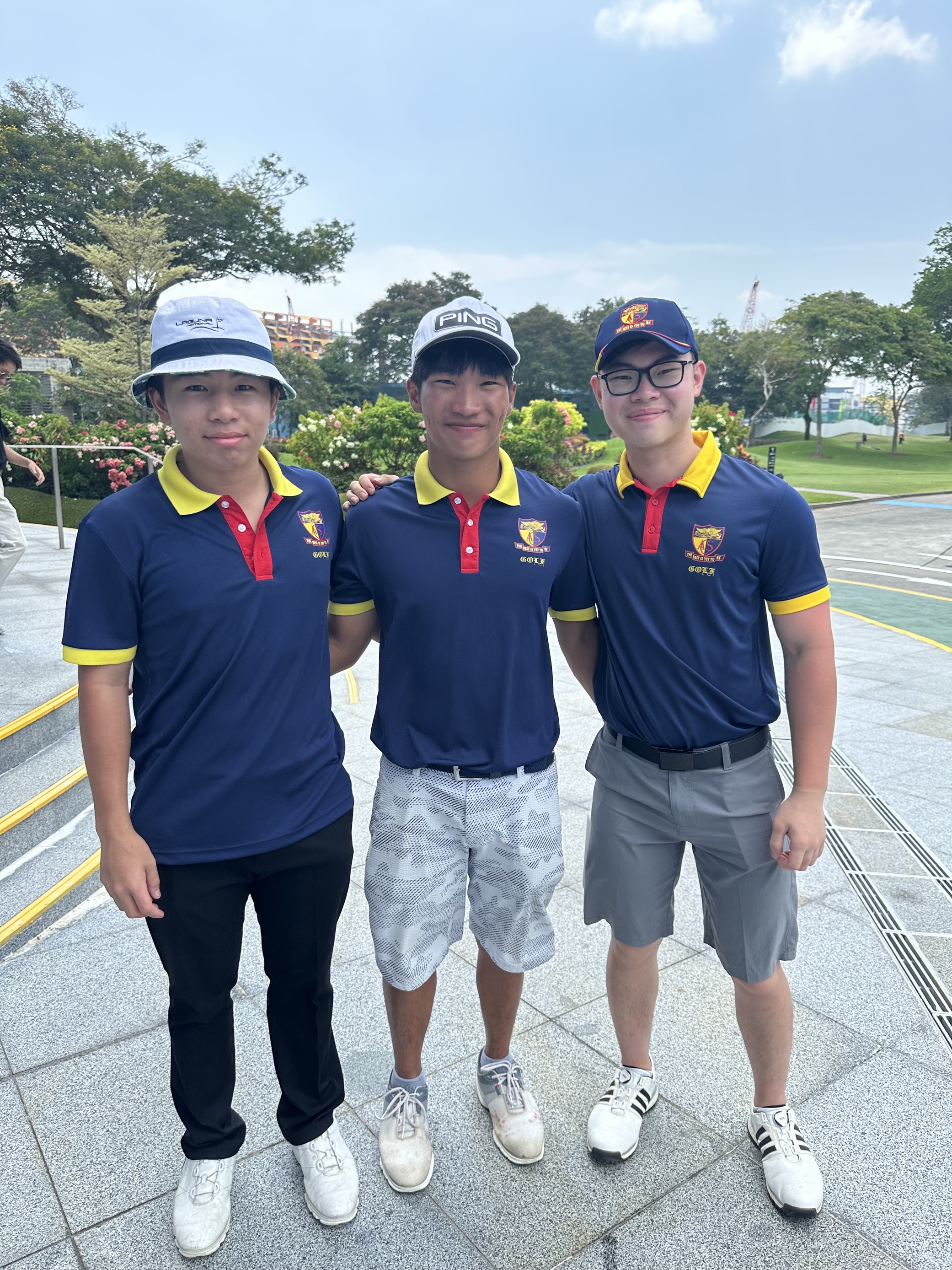 Three young male golfers in matching navy and yellow team polo shirts pose together outdoors at a golf course.